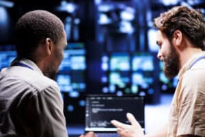 IT developer collaborating with coworker in data center, crafting script code on laptop terminals. Proficient programmers engaging in maintenance work, ensuring optimal functionality and efficiency
