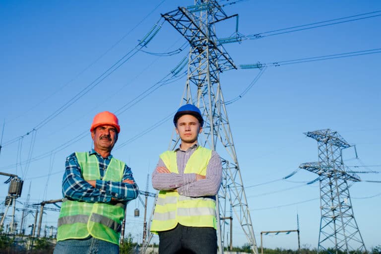 Low angle of confident serious professional male electricians technicians in hardhats and waistcoats looking away while standing together against blue sky and modern electric towers