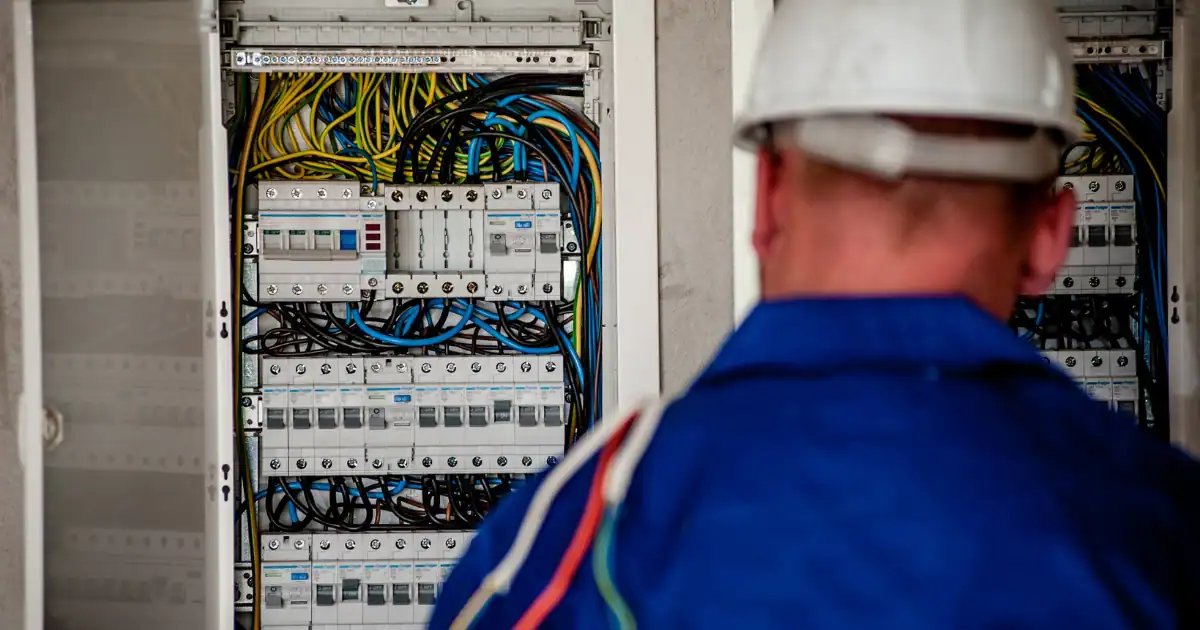 An electrician in a white hard hat and blue coveralls works on an open electrical panel, a task often considered when calculating fire alarm installation costs.