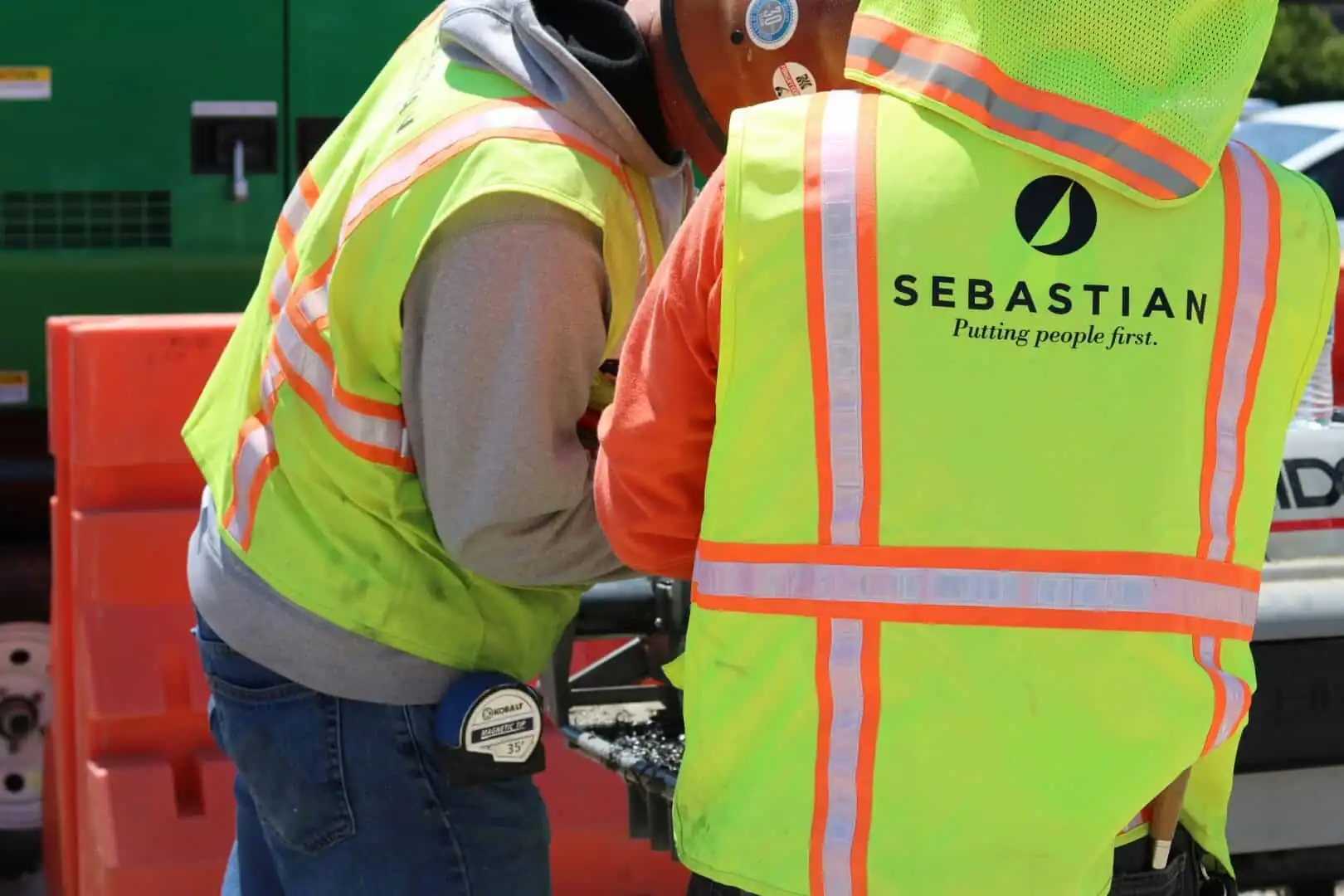 Two industrial electrical contractor workers in high-visibility safety vests at a site, with the Sebastian company logo and slogan visible on the back of one vest.