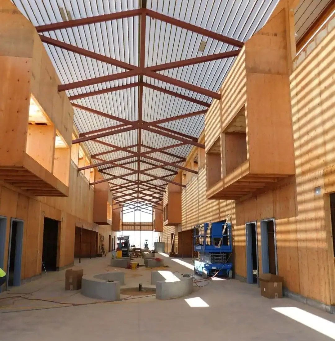 Interior of a building under construction featuring high ceilings with metal trusses, wooden wall panels, and a blue scissor lift on a concrete floor.